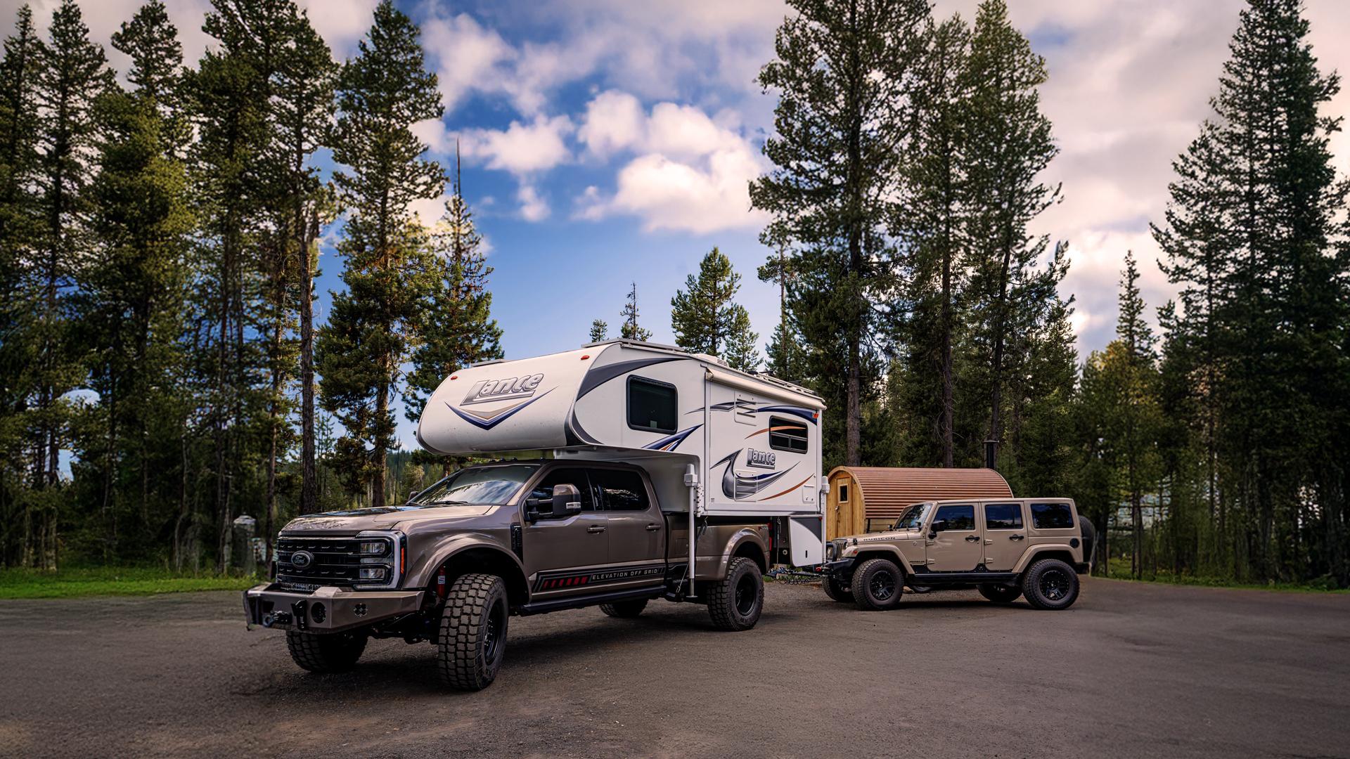 Truck with camper on top in front of pine trees with a jeep behind it. 